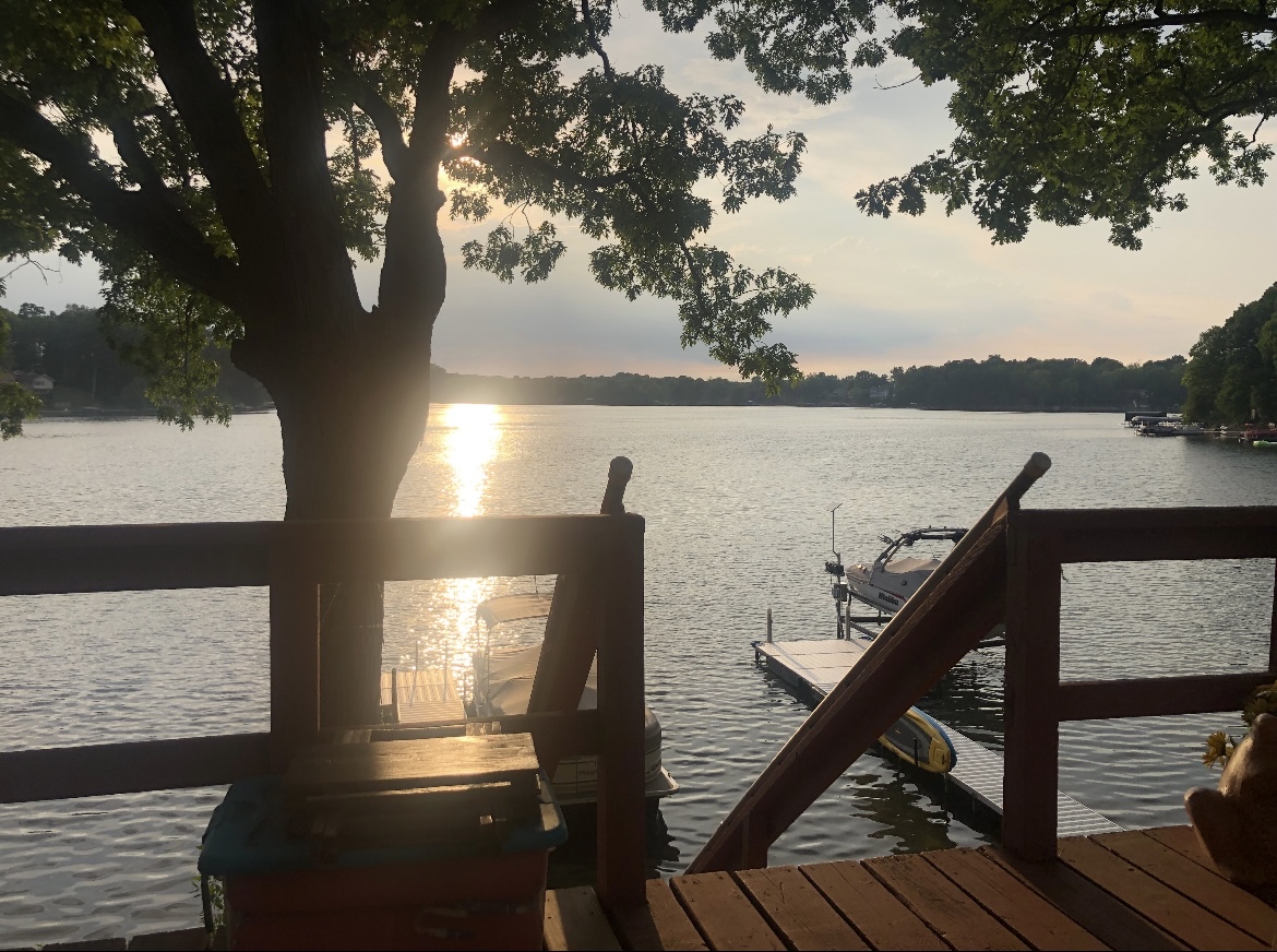 Sunset over the lake seen from the wooden deck, with a boat and dock below and an oak framing the view