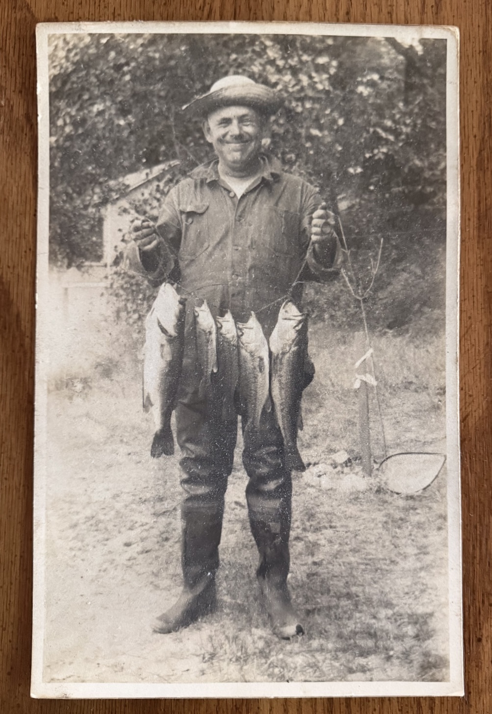 Aged black and white photograph of a man in a straw hat and waders holding a stringer of bass